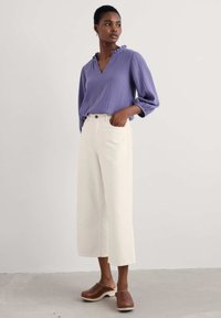 Blusa viola chiaro, testurizzata, con scollo a v e colletto arricciato; abbinata a pantaloni bianchi a vita alta e gamba larga e zoccoli in legno marroni.