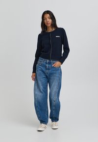 Cárdigan navy de punto con botones y el logo bordado "BALL", combinado con unos jeans anchos de color azul claro y zapatillas blancas. Textura suave y estilo casual.