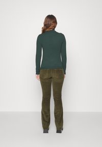 Žena s dlouhými hnědými vlasy, oblečená v tmavozeleném roláku a olivově zelených manšestrových kalhotách, stojící čelem k bílé zdi.