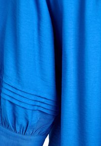 Sinine kangas, millel on kogutud varrukad ja tekstuuriga triibuline muster külje peal, näidates sujuvat tekstuuri ja erksat värvi.