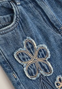 Los jeans de mezclilla presentan una tela azul con una textura desvaída y un distintivo bordado de flores en hilo crema y oscuro en el área del bolsillo delantero.