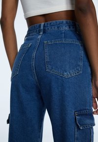 Blå denim cargo byxor med lapplåda, avslappnad passform och synliga söm detaljer. Tyget verkar slätt med en medelviktsstruktur.