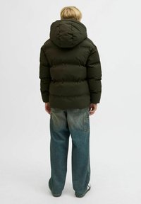 Blandet barn står med ryggen til iført en mørkegrøn dunjakke med hætte og posede, falmede blå jeans på hvid baggrund.