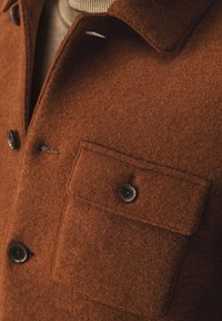 Bruine wollen jas met een kraag, twee zwarte knopen en een patchzak. De stof heeft een zachte en fuzzy textuur.