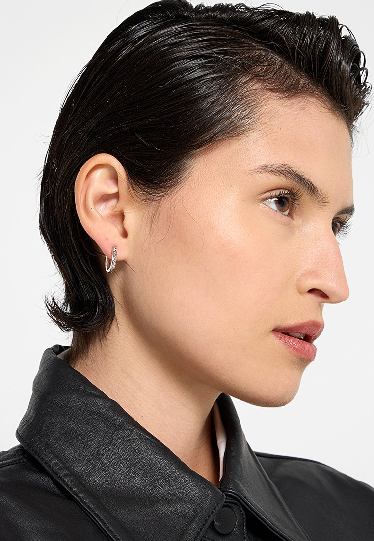 Silberne Creolen mit einem geschwungenen Design und einer strukturierten Oberfläche. Tragen Sie sie zu kurzen Haaren, kombiniert mit einer schwarzen Lederjacke.