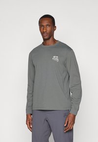 Camisa de manga longa em tecido cinza escuro com um logótipo gráfico branco no lado esquerdo do peito e um corte descontraído. Textura suave e design simples.