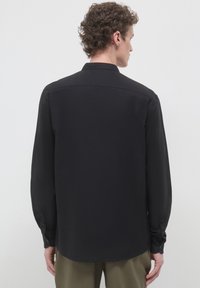 Uomo con capelli ricci che indossa una camicia nera a maniche lunghe e pantaloni verde oliva, in piedi di spalle contro uno sfondo chiaro e uniforme.