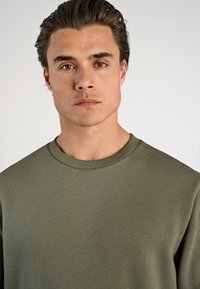 Sudadera verde oliva con cuello redondo, tela texturizada y mangas largas. Diseño simple sin logotipos ni patrones visibles.