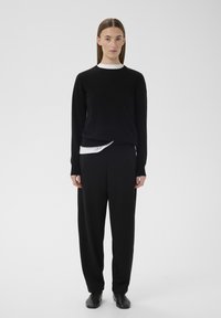 Črn pleten pulover, oblečen čez belo srajco, kombiniran s črnimi ohlapnimi hlačami in črnimi čevlji. Gladek tekstura, minimalističen dizajn.