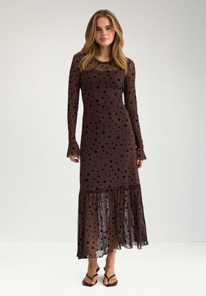 Femme portant une longue robe noire transparente à pois avec un ourlet à volants et des sandales noires, debout devant un fond clair uni.