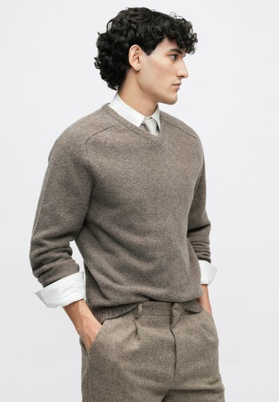 Maglione marrone in tessuto morbido e testurizzato con scollatura a V, indossato sopra una camicia bianca con colletto, abbinato a pantaloni grigi a spina di pesce.