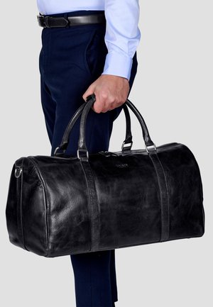Black leather duffel bag with two sturdy handles, subtle sheen, and light stitching details, carried by a person in navy pants.