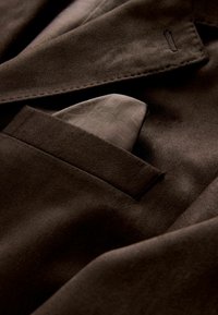 Hnědý sametový blazer s hladkou texturou a kapesním čtvercem viditelným v náprsní kapse, který má jemný vzor.