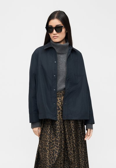 Γυναίκα με μαύρα γυαλιά ηλίου, ναυτικό μπουφάν με κουμπιά, γκρι πουλόβερ με ζιβάγκο και φούστα με λεοπάρ μοτίβο σε απλό λευκό φόντο.