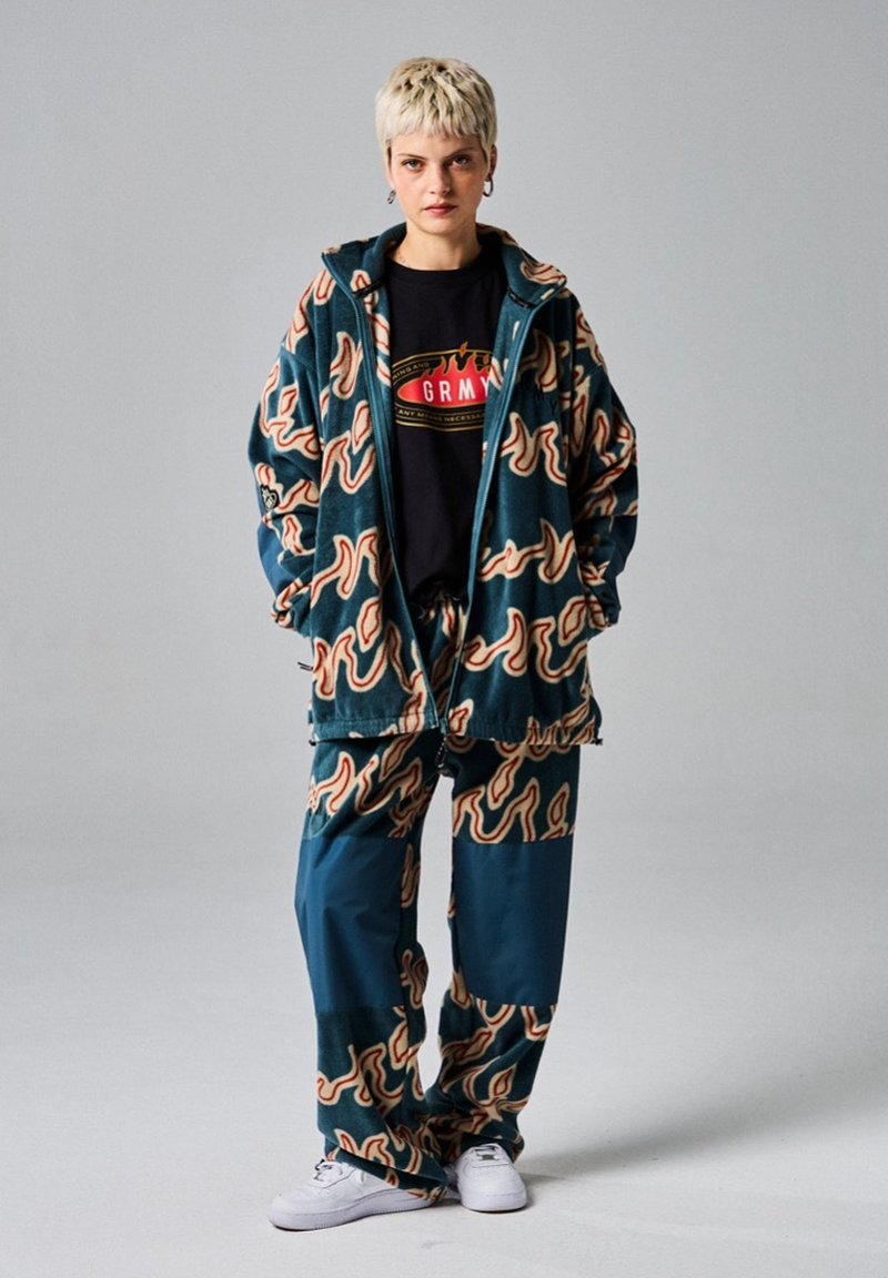 Fleece'owa kurtka z kapturem o zielonym tle, z abstrakcyjnym brązowym i kremowym wzorem; pasujące spodnie; czarna koszulka z logo; białe trampki.