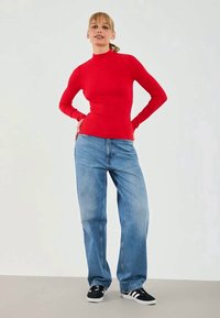 Suéter de gola alta vermelho com textura em relevos, emparelhado com jeans de lavagem clara e corte solto, e sapatilhas atléticas pretas com riscas brancas.