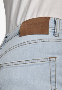 Bolsillo trasero de vaqueros de mezclilla azul claro con una etiqueta de cuero marrón cosida arriba, con el texto "DAN JOHN DENIM" grabado en la etiqueta.