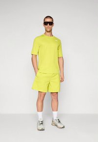 Lime zöld sportpóló és rövidnadrág könnyű anyagból; a modellen sötét napszemüveg és szürke, fekete részletekkel ellátott sportcipő látható.