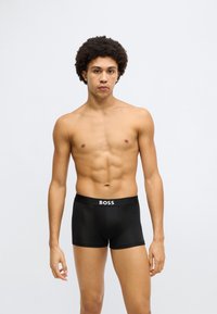 Fekete boxeralsók puha anyagból, rugalmas derékpánttal, amelyen fehér „BOSS” felirat látható elöl. A modell vékony testalkatú.