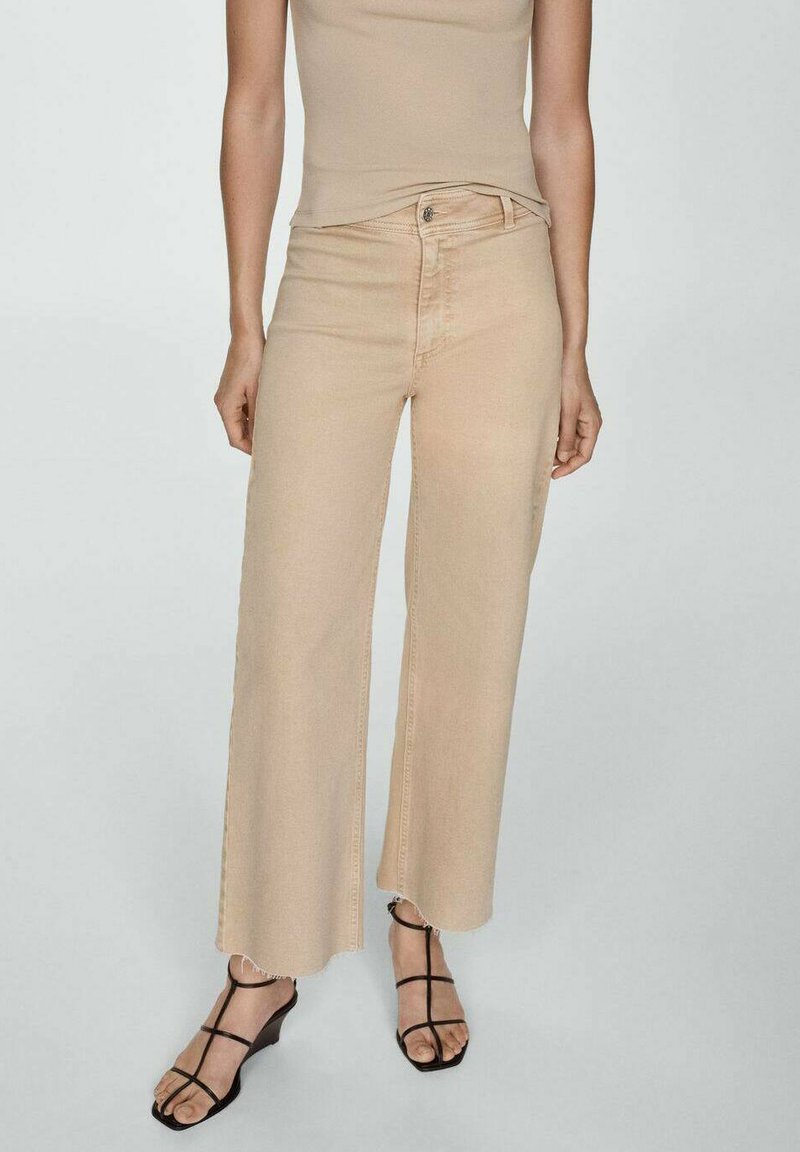 Beige wide-leg jeans feature a button and zipper closure, raw hem, and soft denim texture; paired with black strappy sandals.