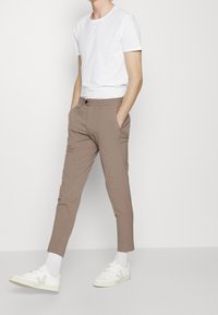 Pantaloni beige a gamba affusolata realizzati in tessuto leggero, dotati di chiusura frontale con bottone e tasche laterali, abbinati a una t-shirt bianca semplice e sneakers.