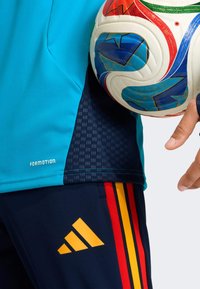 Urheiluasu, jossa turkoosi paita, jossa teksturoidut laivastonsiniset sivupaneelit ja sinisiä tehosteita. Tummat housut, joissa punaiset ja keltaiset raidat, Adidas-logo näkyvissä.