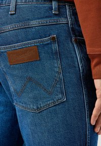 Jeans de mezclilla azul oscuro con un parche de cuero marrón que lleva el texto "Wrangler", acentuados por un patrón de costura en zigzag en el bolsillo trasero.