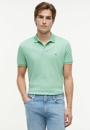 Junger Mann, der ein hellgrünes Poloshirt und hellblaue Jeans trägt, mit einer Hand in der Tasche vor einem schlichten weißen Hintergrund steht.