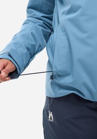 Light blue waterproof jacket with adjustable hem. Features textured fabric and pull cord on the side. Dark pants with logo detail.