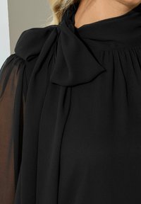 Blusa negra con cuello alto y detalle de lazo, que presenta mangas largas transparentes y una tela texturizada. Primer plano del cuello y la textura de la tela.