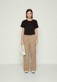 Donna che indossa una t-shirt nera, pantaloni cargo beige a gamba larga, scarpe bianche aperte sulla punta, mentre tiene una borsa bianca, in piedi davanti a uno sfondo chiaro e semplice.
