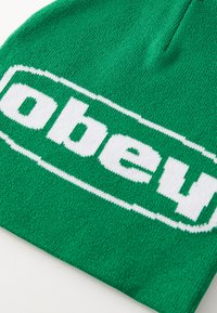 Zaļš adīts beanie ar izteiksmīgu balto "obey" logotipu. Tekstūra ir mīksta un elastīga, ar bezšuvju dizainu un kārtīgu apdari.