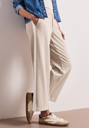 Osoba nosící béžové široké kalhoty s bočními pruhy, hnědé a krémové tenisky, bílý top a modrou džínovou bundu s rukama v kapsách.