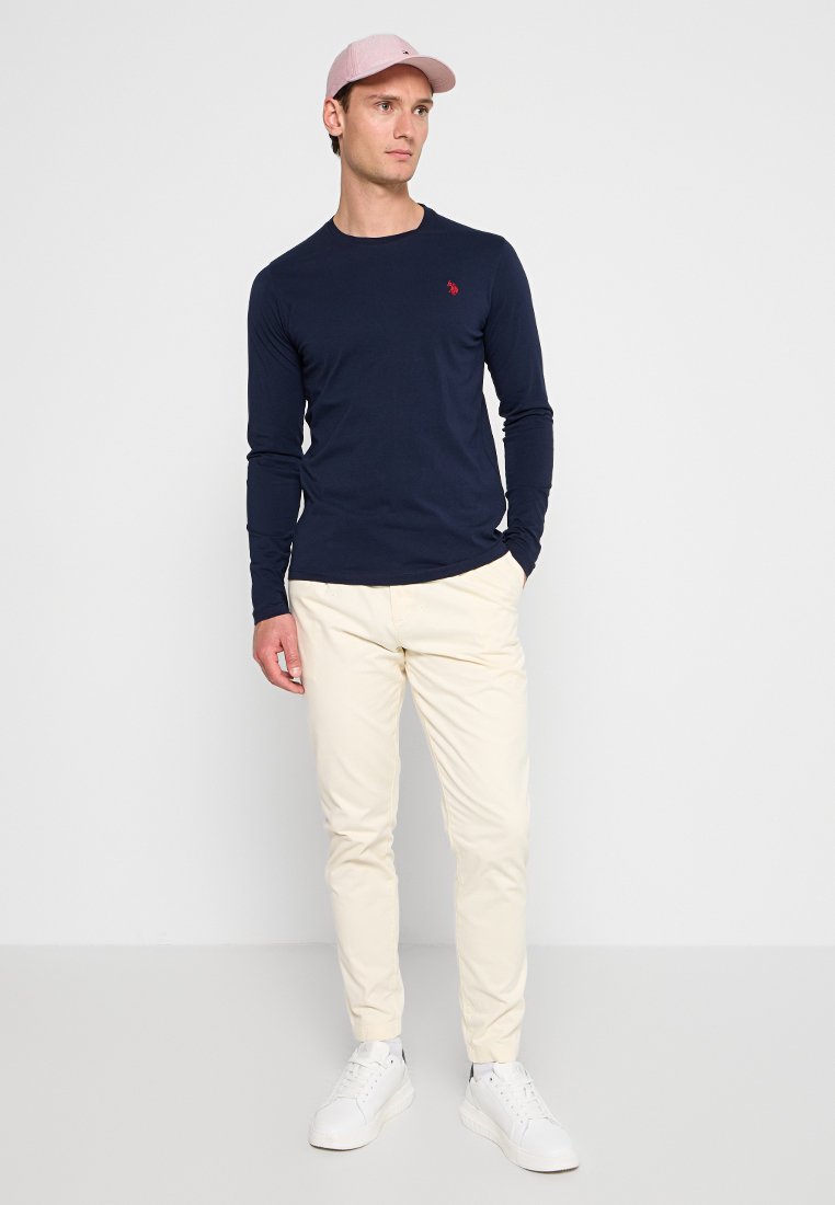 Marineblaue Langarmbluse mit kleinem roten Logo, kombiniert mit hellbeigen Slim-Fit-Hosen und weißen Sneakers; trägt eine pinke Kappe.