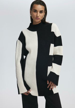 Maglione over-size a costine con pannelli alternativi neri e crema, colletto alto e maniche ampie con una vestibilità rilassata. Dettagli neri presenti.
