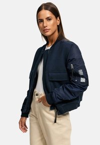 Navy bomber jacket with a mix of textured fabrics, zippered pockets, and a smooth satin sleeve. Worn over a white shirt and beige pants.