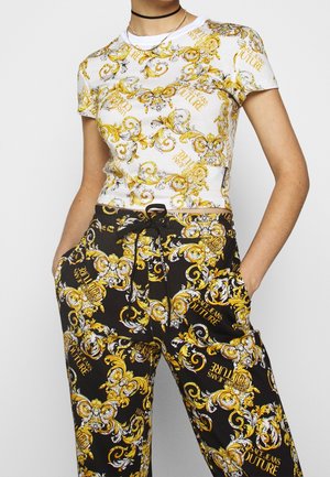 Witte t-shirt met gele en zwarte barokprint; zwarte broek met een vergelijkbaar patroon, voorzien van een koord en zakken.