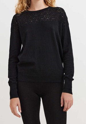 Donna con maglione nero a maniche lunghe con motivo a diamante testurizzato sulla parte superiore del petto e sulle spalle, abbinato a pantaloni neri.