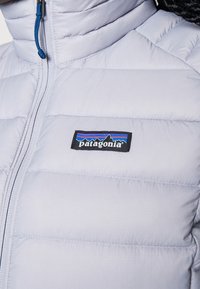 Jasnoszara kurtka puchowa z pionowym pikowaniem, pełnym zamkiem błyskawicznym i czarnym logo Patagonia na piersi. Posiada niebieski uchwyt zamka.