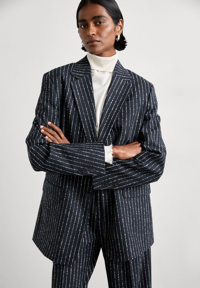 Ναυτικό μπλέιζερ με ρίγες, σε κουτί εφαρμογή, με κεντρικά περάσματα και μπροστινές τσέπες. Διαθέτει υφή υφάσματος με λευκές κάθετες γραμμές.