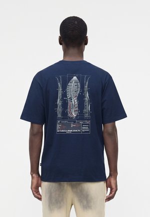 Homme portant un t-shirt bleu marine avec un design détaillé blanc et rouge d’un plan de semelle de chaussure dans le dos.