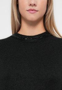Pull noir à corps texturé et col orné de paillettes noires brillantes. Gros plan mettant en valeur l'encolure et le matériau.