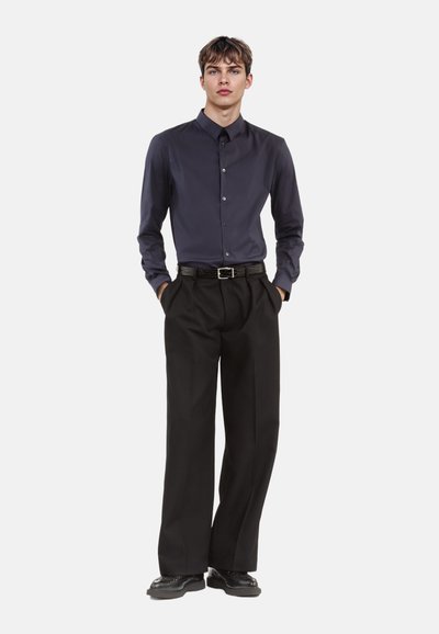 Donkerblauwe button-up shirt met een kraag, gecombineerd met zwarte wijde broek en een gestructureerde riem. De outfit heeft gepolijste zwarte schoenen.