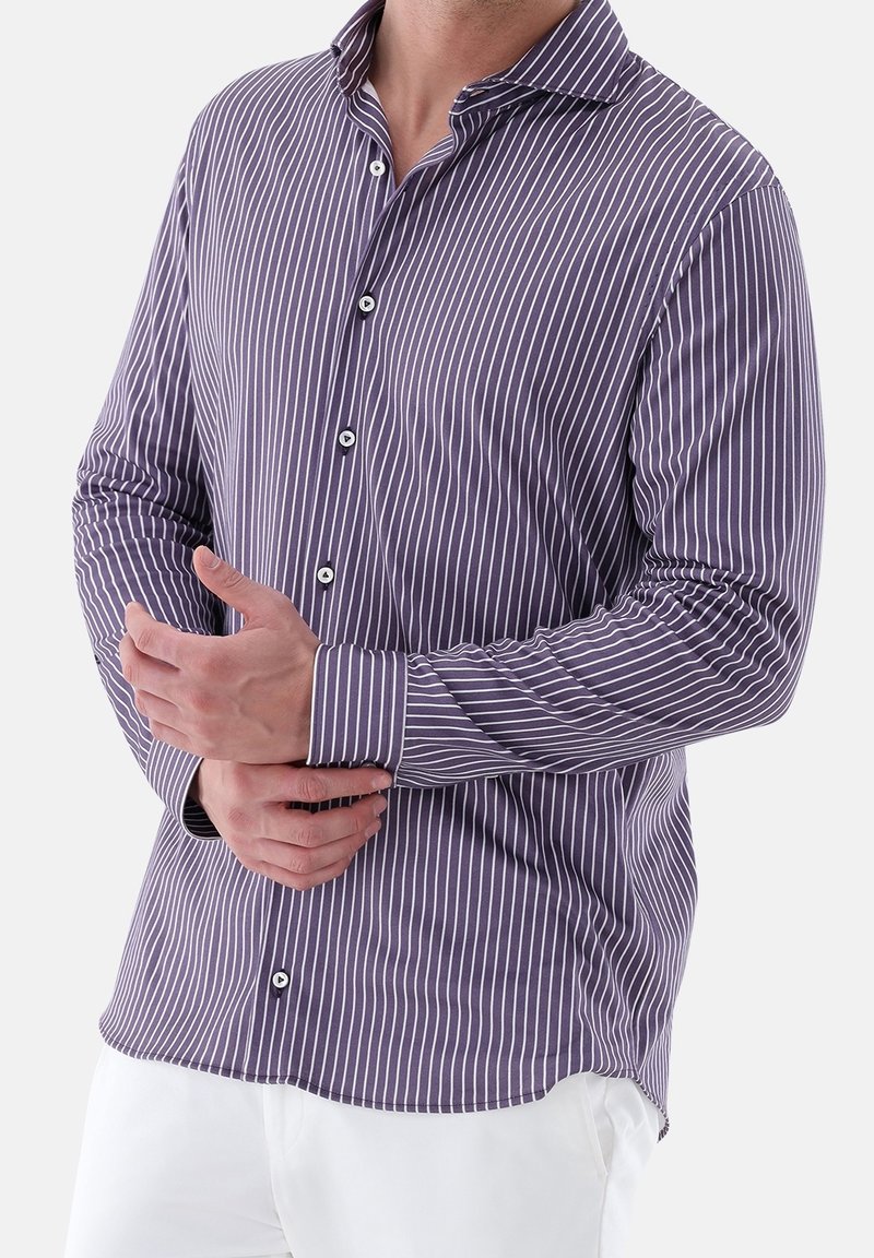 Homme portant une chemise violette boutonnée avec des rayures blanches verticales, ajustant le poignet, assortie à un pantalon blanc.