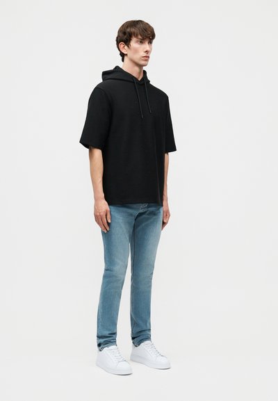 Νεαρός άνδρας στέκεται με το πρόσωπο προς τα δεξιά, φοράει μαύρο φούτερ με κοντά μανίκια, ανοιχτό μπλε τζιν και λευκά αθλητικά παπούτσια σε απλό γκρι φόντο.