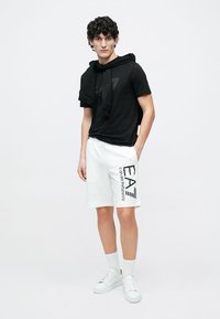 Sort T-skjorte med kortermet hette, kombinert med hvite shorts med en stor logo på siden og hvite joggesko.