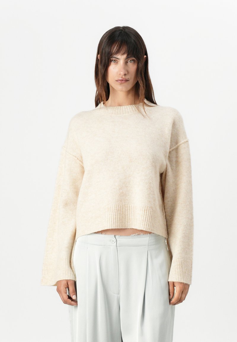 Beige cropped sweater with a textured finish, ribbed cuffs, and a round neckline, paired with light gray, pleated pants.
