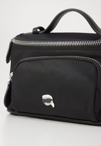 Black nylon bag with silver zipper, front pocket, and a round pin detail featuring a character in sunglasses. Structured shape, top handle.