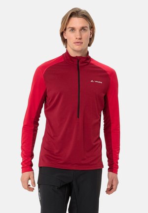 Homme blond portant un pull de sport rouge à manches longues avec une demi-fermeture éclair noire et un pantalon noir, debout sur un fond blanc.