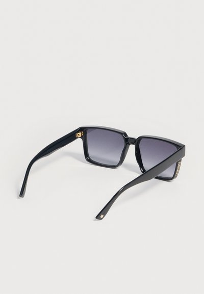 Μαύρα ορθογώνια γυαλιά ηλίου με σκούρους φακούς gradient και χρυσές λεπτομέρειες στις μεντεσέδες και τα άκρα των βραχιόνων, τοποθετημένα σε λευκή επιφάνεια.